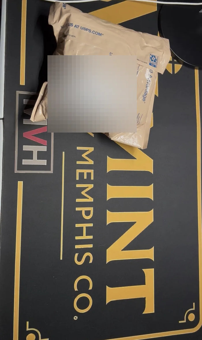 A padded USPS mailer from an inbound collection resting on the Mint By Memphis mat, its shipping label blurred to protect the seller's privacy.